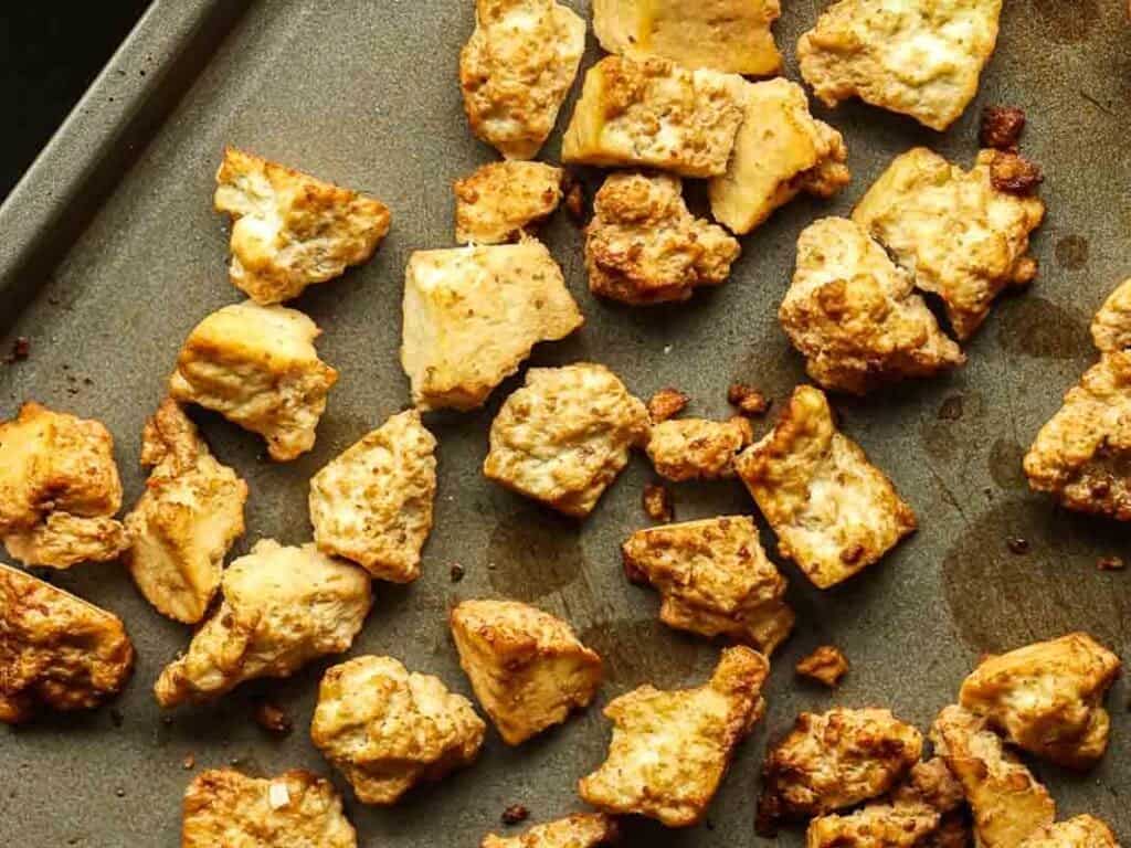 Golden-brown roasted tofu pieces scattered on a dark baking sheet, displaying a crispy texture and lightly charred edges.